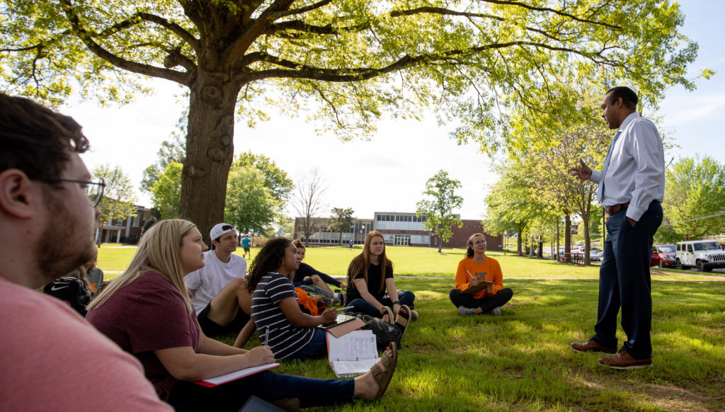 A professor lectures outdoors for a group of students sitting in the shade of a tree