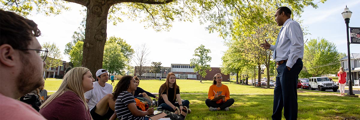 Professor gives a lecture outside to students sitting on grass underneath a tree.