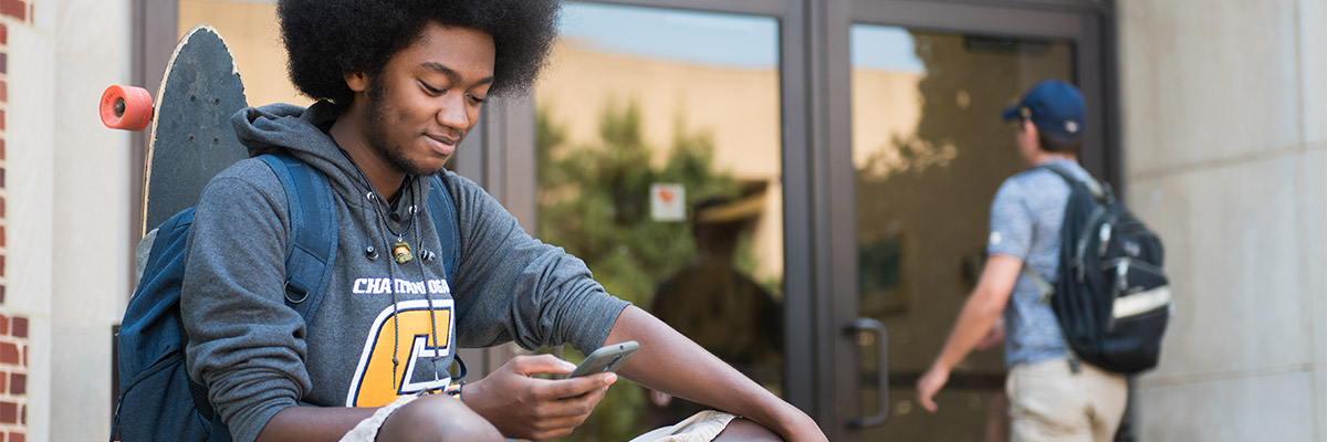 Black student wearing UTC hoodie sits while using his phone.