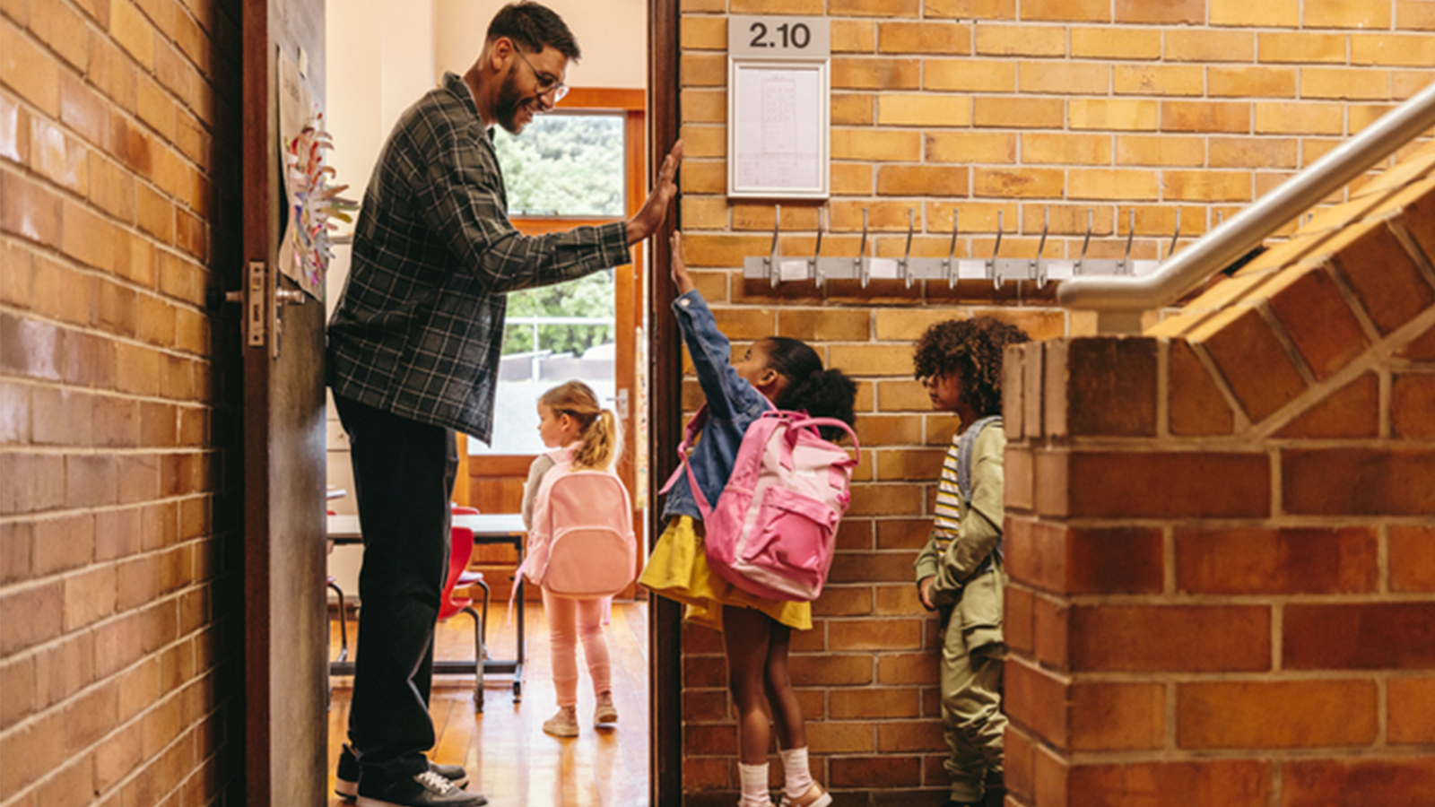 Stock photo of Elementary school teacher greeting his students at the door. Male teacher welcoming his class with a high five outside their classroom. Child mentor motivates his pupils in a primary school.