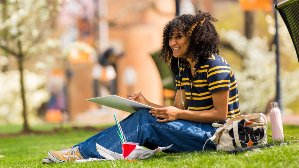 College student smiling while doing class work on a campus lawn.