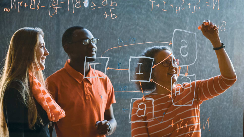 2 people look on while a third writes equations on a glass surface