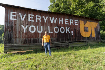 Man in orange polo shirt standing in front of an old barn, with the words "Everywhere You Look, UT" painted on the side.
