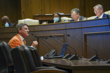 Man in orange business suit speaks into a microphone, in front of a room of state officials.