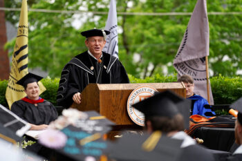 A man garbed in commencement regalia speaks to a crowd of graduating students.
