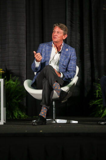 A man in a checkered blue jacket speaks from a stage while seated.