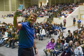 A man in a blue polo takes a selfie with a crowd of students in the stadium behind him.