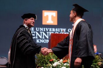 A man in commencement regalie shakes hands with a graduating student as he receives his diploma.