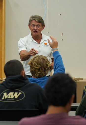 A man in a white polo shirt points to a student with the hand raised for a question.
