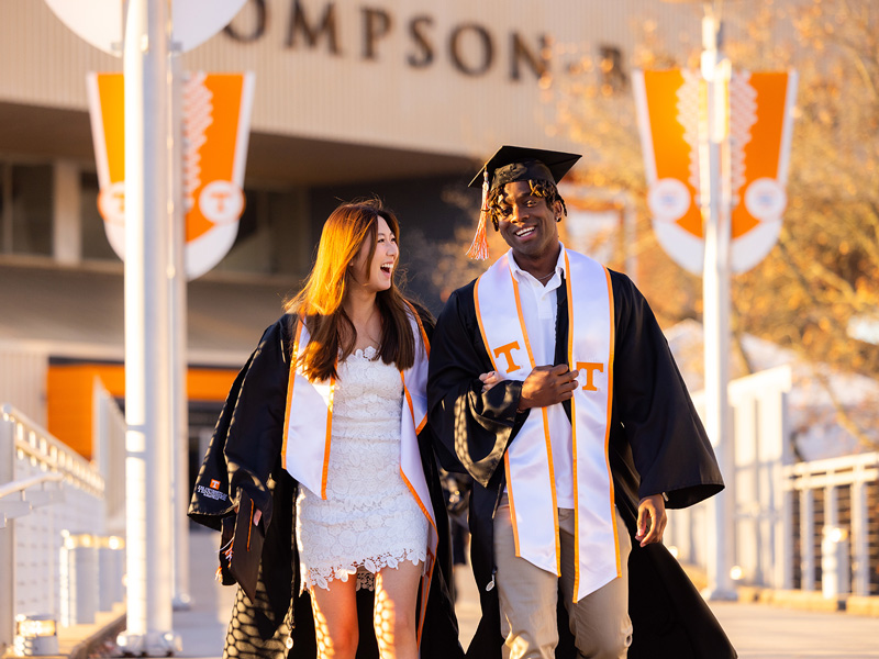 A young female and male student adorned in UT Knoxville colors and graduation robes walk across campus.
