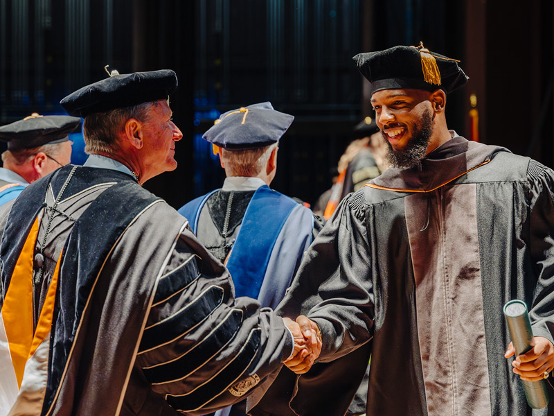 A black student with a beard shakes hands with UT System President Randy Boyd during a commencement ceremony.