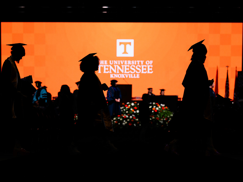 Silhouettes of three students walking across the stage at commencement, with a bright orange backdrop in the background.
