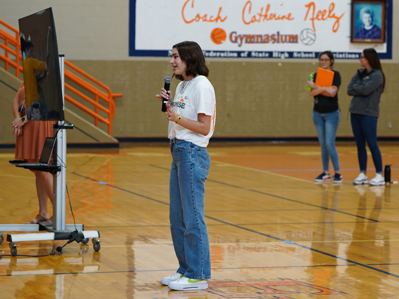 A young college student, wearing a white t-shirt and blue jeans, standing in a gymnasium speaking into a microphone.