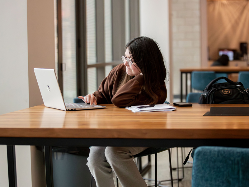 A young woman with brown hair and glasses leans on a table while working on her laptop.