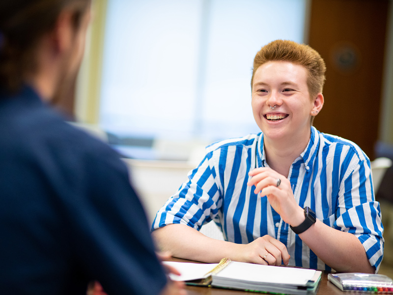A young college student with short red hair and a white-blue striped shirt talks with a counselor.