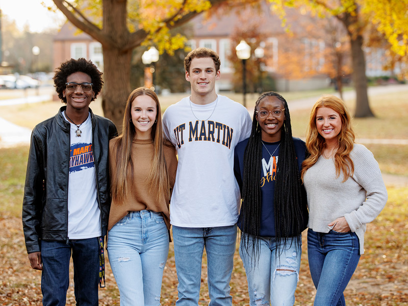 A diverse group of two young men and three young women stand shoulder to shoulder for a group photo.