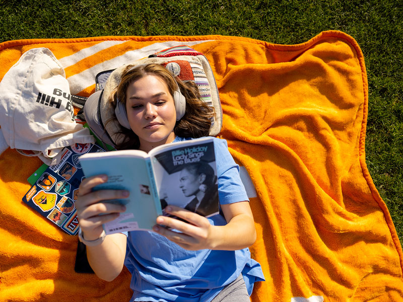 A female college student lays on an orange blanket on the ground outside, reading a book.