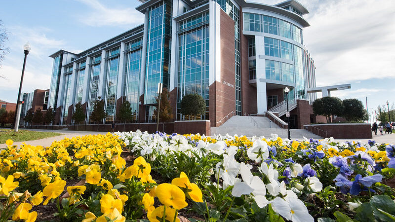 Scenic photo of a red brick and glass building, with yellow, white, and blue flowers in front of the building.