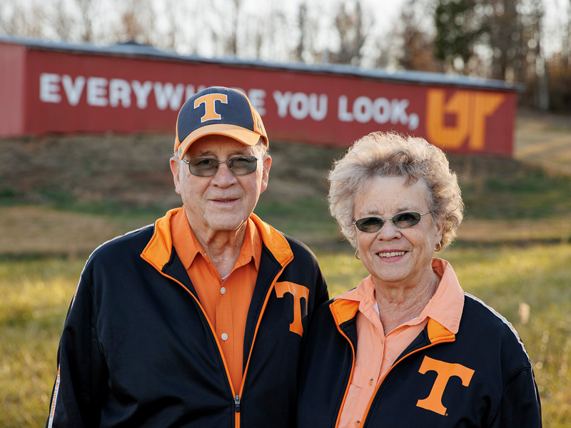 An elderly couple, both wearing black jackets with the UT Knoxville Power T embroidered on it, stand in a field in front of a red barn.