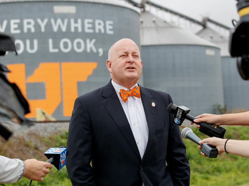 A man with a black jacket, white shirt and orange bowtie talks to reporters holding microphones in front of them.