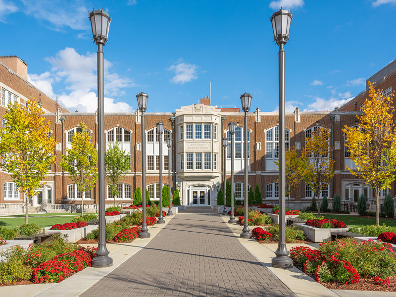 Red brick administration building with a row of light poles lining a pathway, surrounded by red flowers and blue skies.