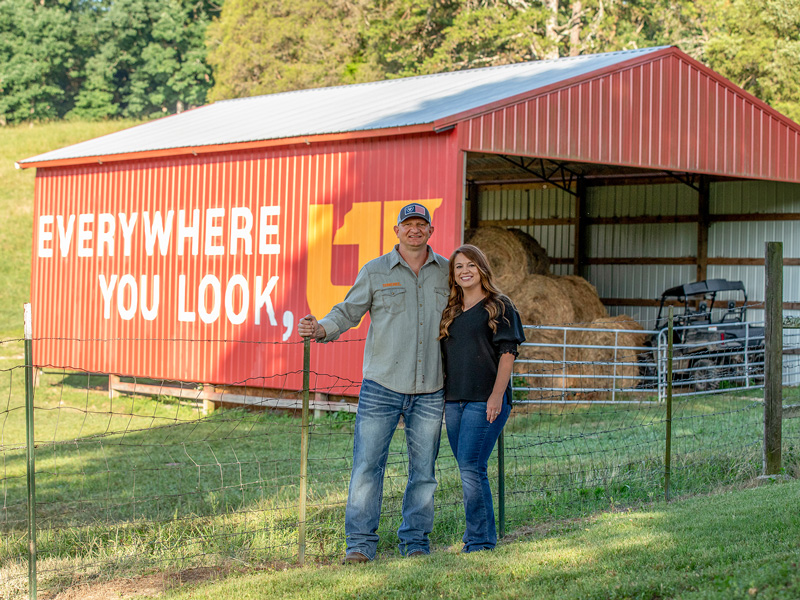 A couple standing in front of their red barn with the words "Everywhere You Look, UT" painted on the side.