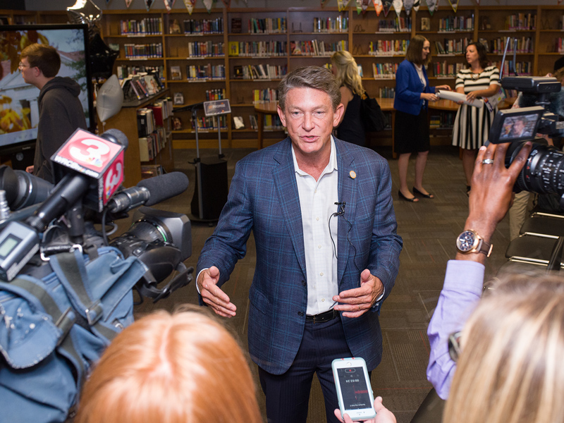 A man in a blue suit speaks to a group of reporters with microphones and cameras while inside of a library.