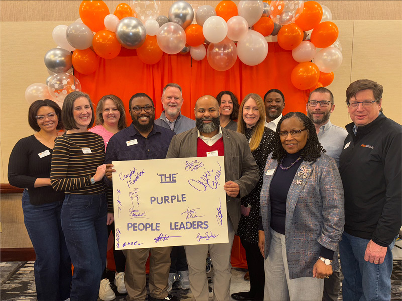 A group of people pose for a photo against an orange backdrop decorated with white and orange balloons.