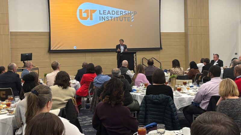 A room full of people sitting around tables listen to a man speaking from behind a podium.