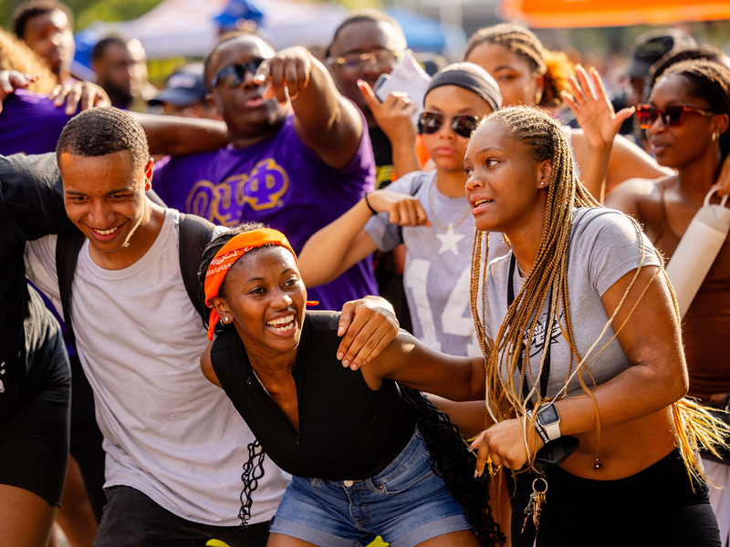 A group of multicultural students dance and sway, arm-in-arm on a campus space.