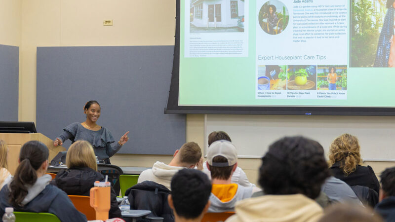 A woman speaks to a room of students while pointing at a presentation screen.