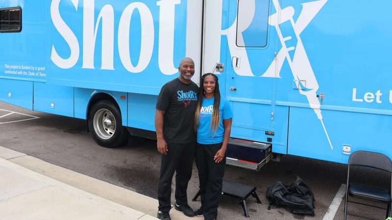 A man and woman standing next to a large, blue trailer with the logo "Shot RX" branded on it.