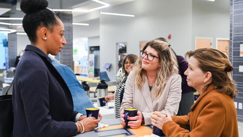 Three women stand around a table with coffee while talking.