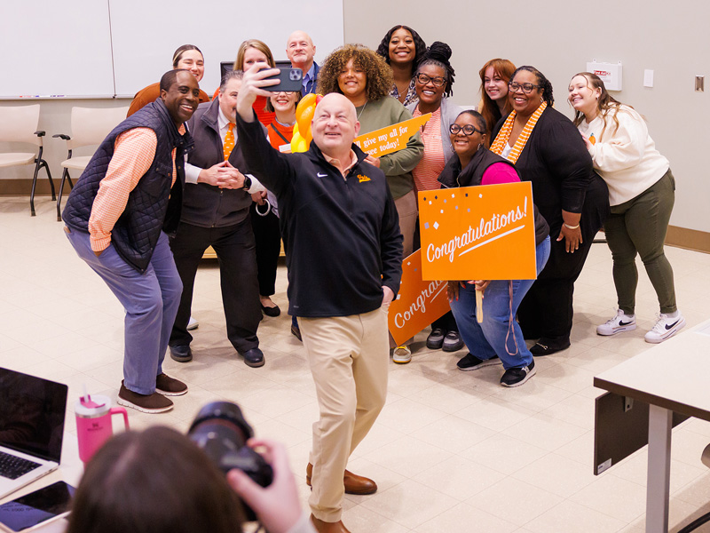 A group of people stand behind a man holding up a cell phone to take a group selfie.