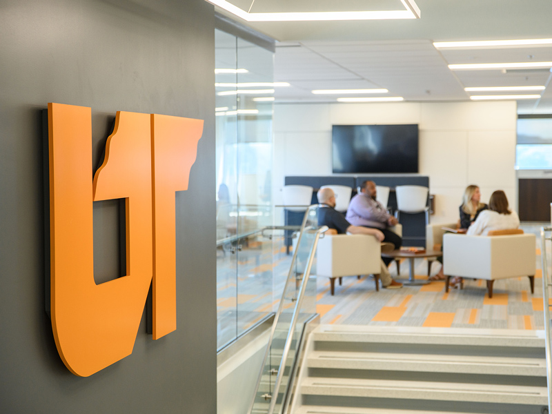 A orange UT icon sign mounted on a wall in the foreground, with a group of people sitting and talking in the background.