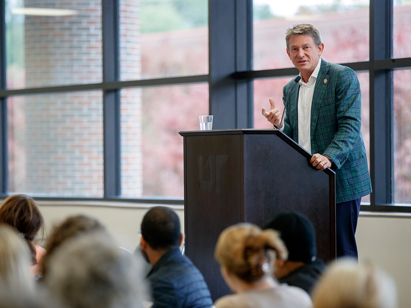 A man in a business suit speaking from behind a podium to a room full of employees.