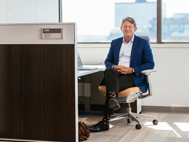 An older man in a blue blazer sits at a cubicle desk next to a window.