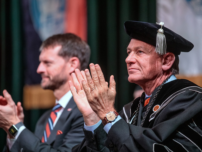 Close-up shot of two men wearing business suits and commencement regalia clapping during a graduation ceremony.