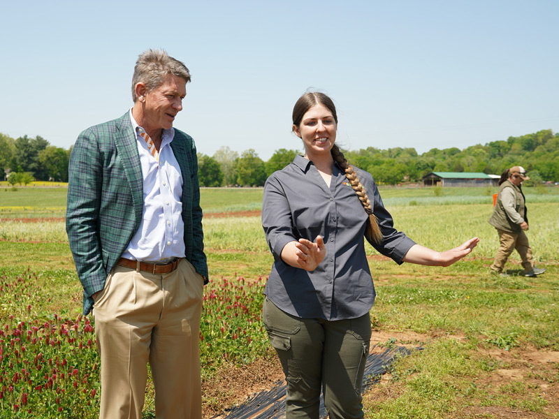 A man in a green blazer listens to a woman presenting something in a large, open field, while another woman walks by in the background.
