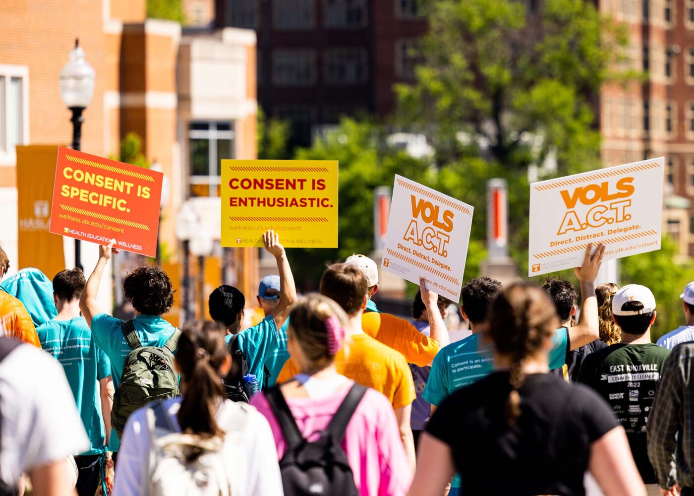 A crowd of students walking, holding multiple red, yellow and white signs themed around consent and Sexual Assault Awareness month.