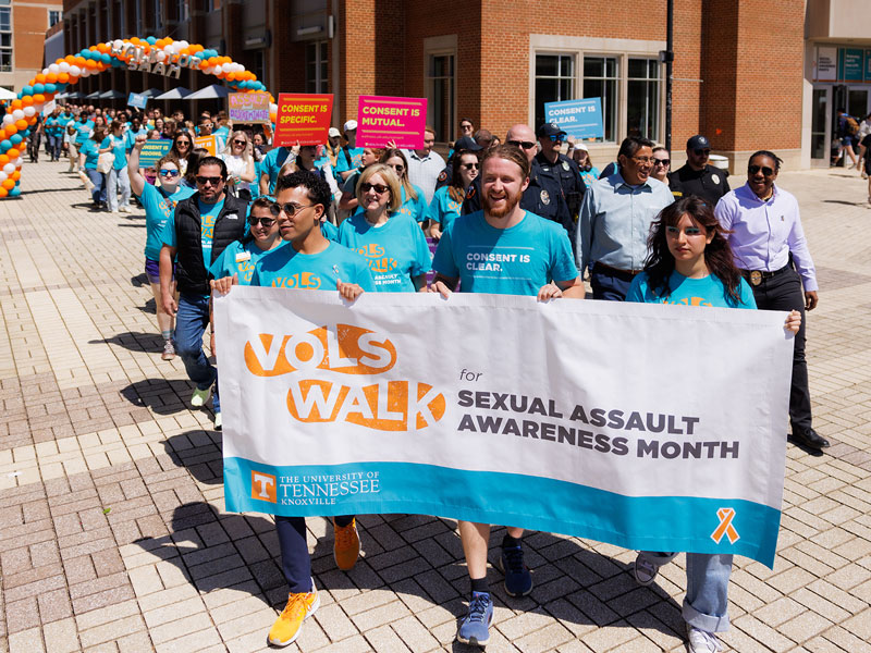 A crowd of people wearing blue shirts walk with a banner saying "Vols Walk for Sexual Assault Awareness Month."