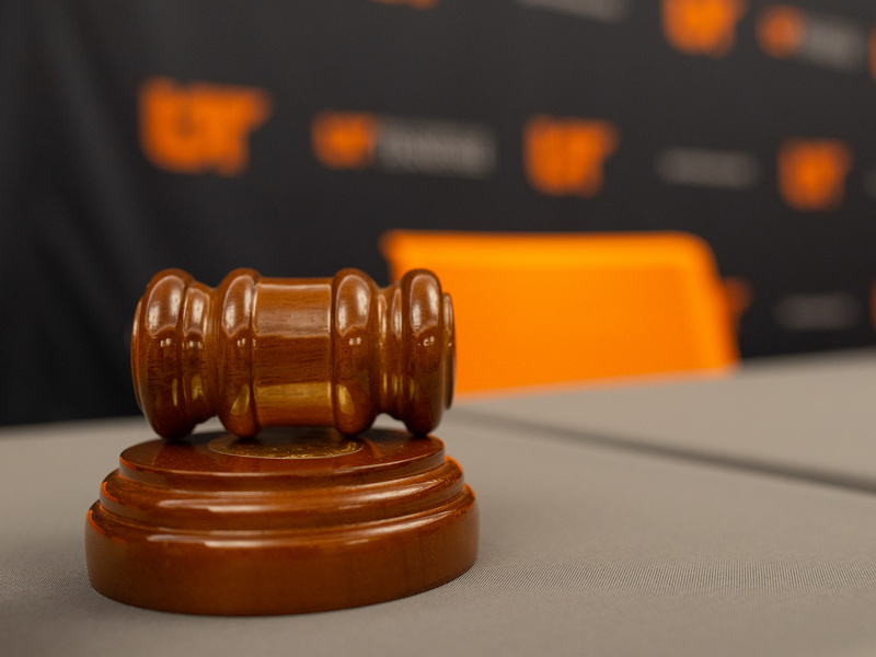 A wooden gavel sitting on table, with a black UT System branded backdrop behind.