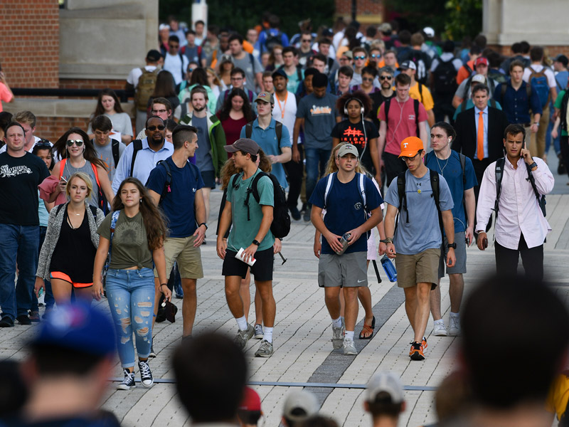 A large, diverse crowd of students walk down a paved pathway on a college campus.