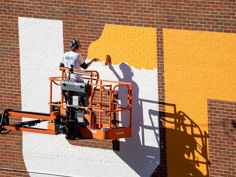 Mural artist painting the UT icon in orange on a brick building.