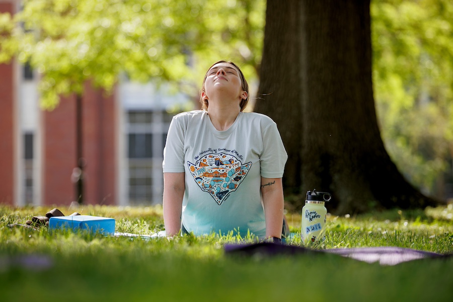 A female student practicing yoga on a grassy campus quad.