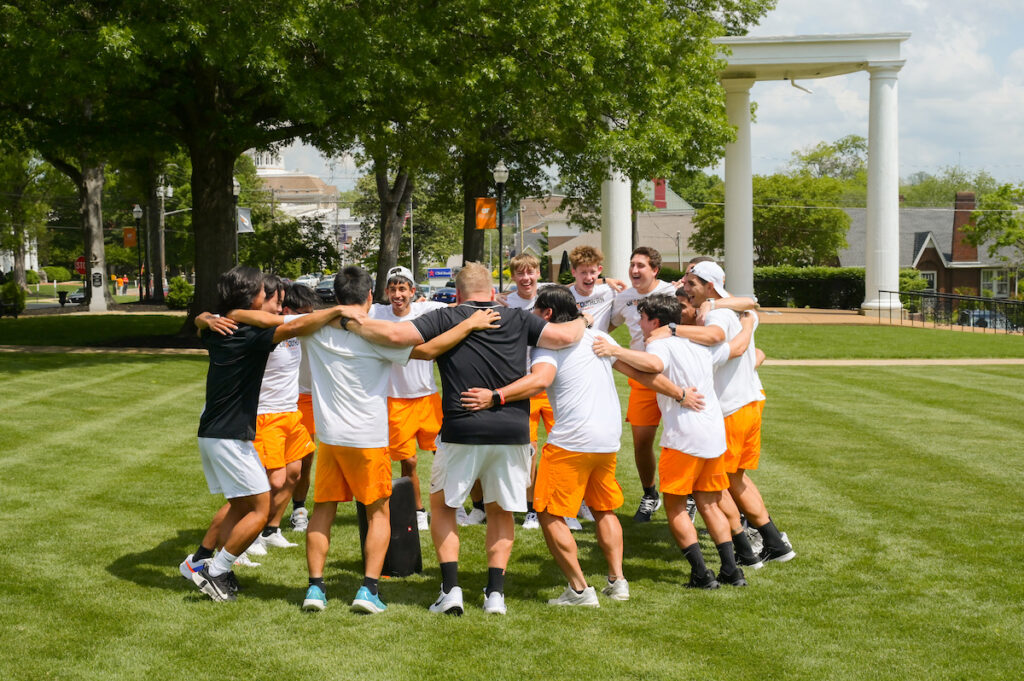 UT Southern men’s tennis team celebrates in a huddle on the campus lawn, wearing white shirts and orange shorts with the iconic white columns in the background.