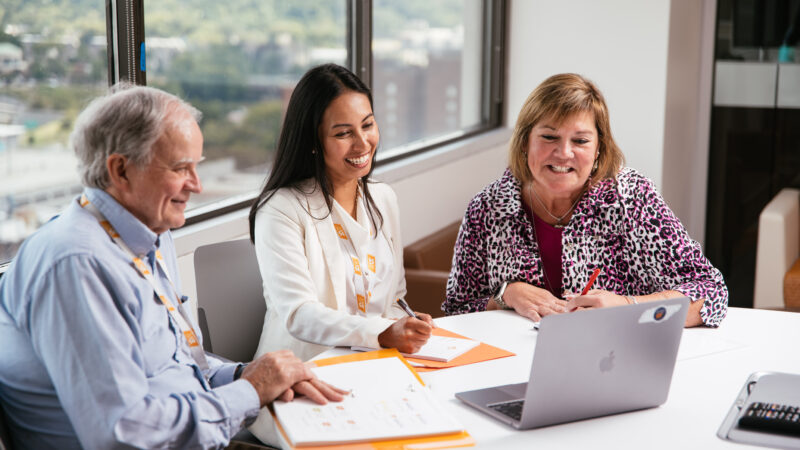 Three UT employees sit at a round table in a brightly lit office, smiling and taking notes during a virtual meeting on a laptop.