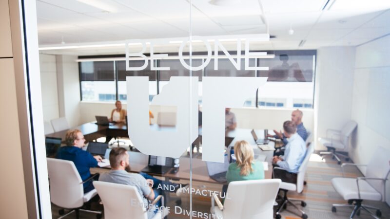 Team meeting in a conference room with the Be One UT values promoted on the glass wall.