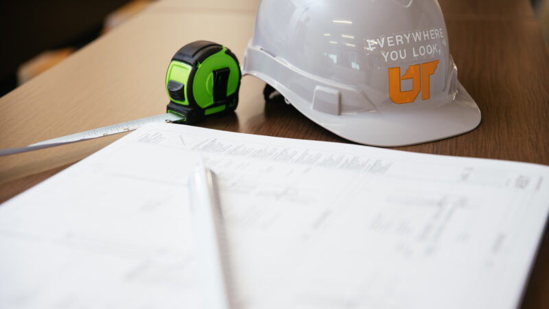 Close-up of a UT-branded hard hat and tape measure on a table beside construction blueprints.