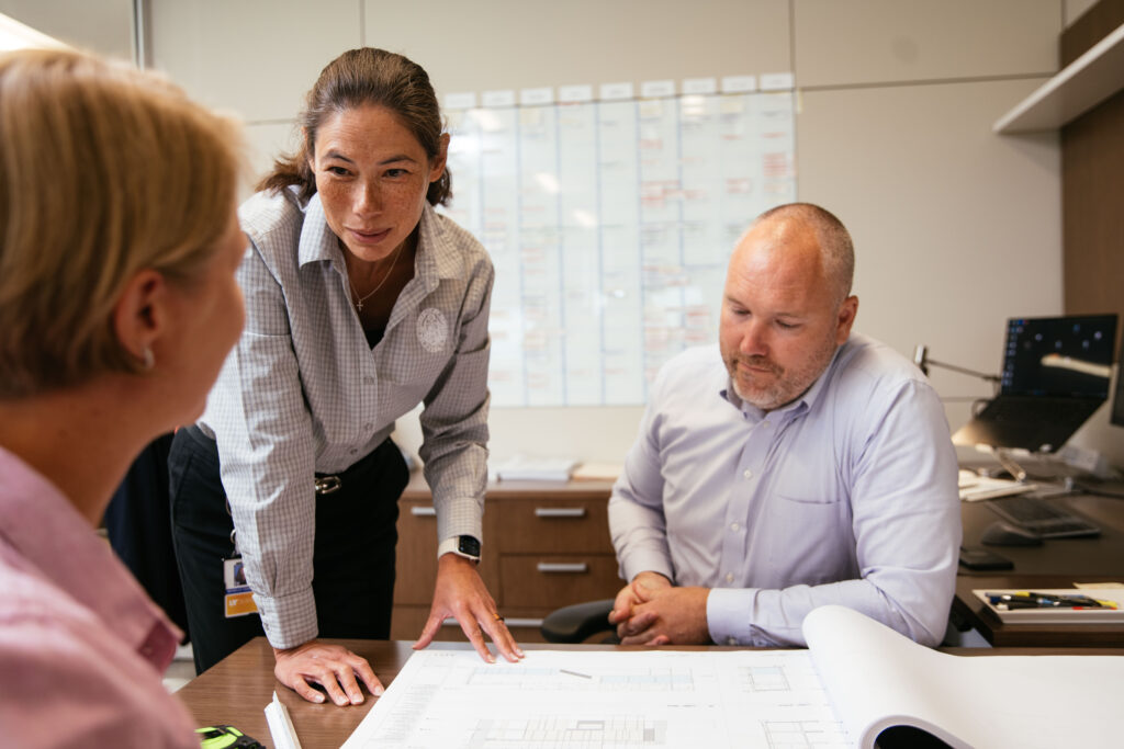 Three UT employees gather around a desk to discuss blueprints spread out on the surface.